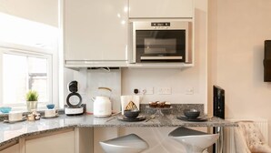 Uma cozinha moderna com armários brancos, forno embutido, chaleira e dois banquetas altas.