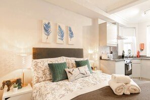 A modern bedroom with a large bed, a nightstand, a lamp, and a view of the kitchen. - 12 Helena House Studio (Reading)
