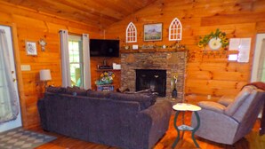 A cozy log cabin interior with a stone fireplace, wooden walls, a sofa, and a chair. - ❤ VERY ROMANTIC CABIN ❤ Near Cades Cove, WIFI, Hot Tub, Jacuzzi, Sauna (Townsend)