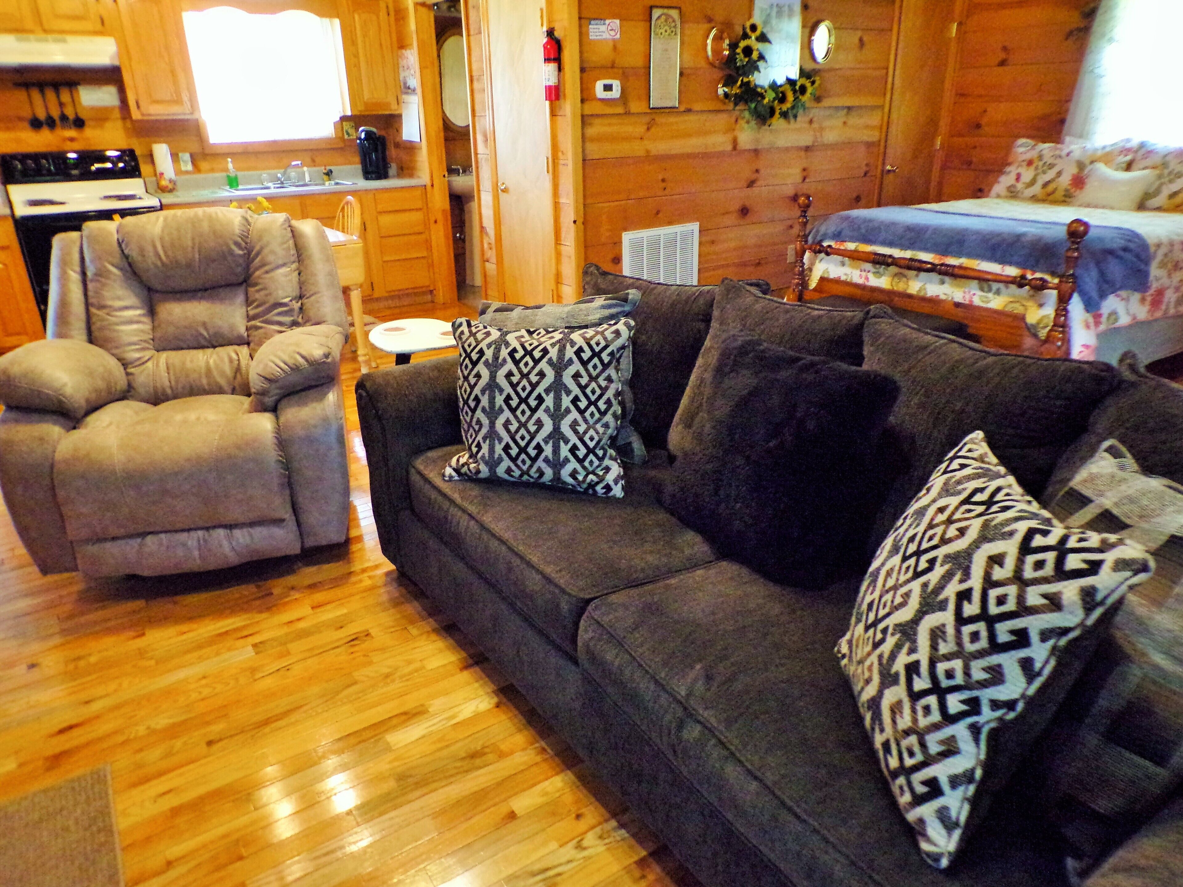 L’intérieur d’un chalet accueillant, avec un plancher en bois, un canapé agrémenté de coussins à motifs, un fauteuil inclinable et un lit avec une literie à fleurs.