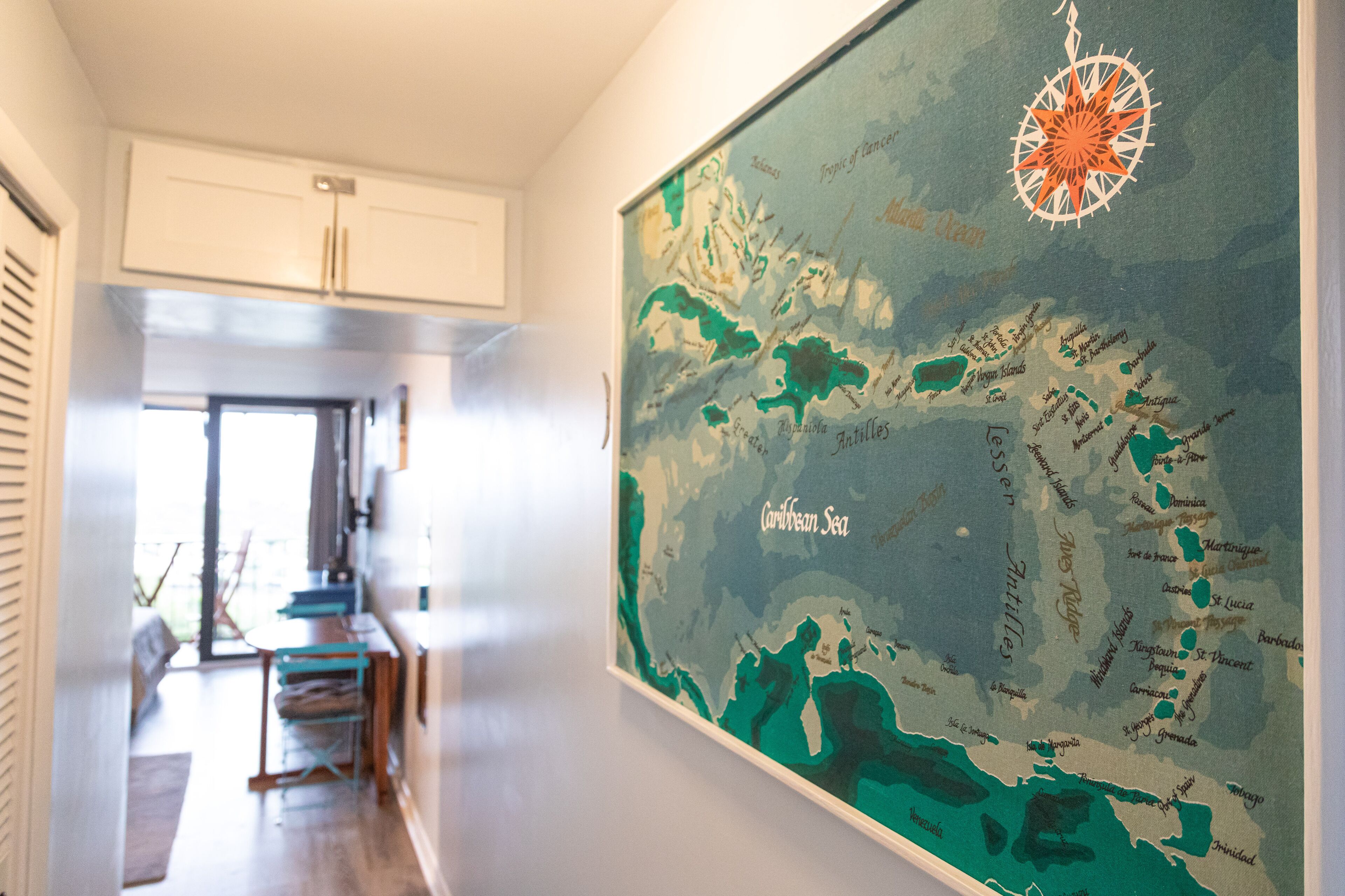 A hallway with a large map of the Caribbean Sea on the wall, a dining area with a table and chairs, and a kitchen area with cabinets and a refrigerator.
