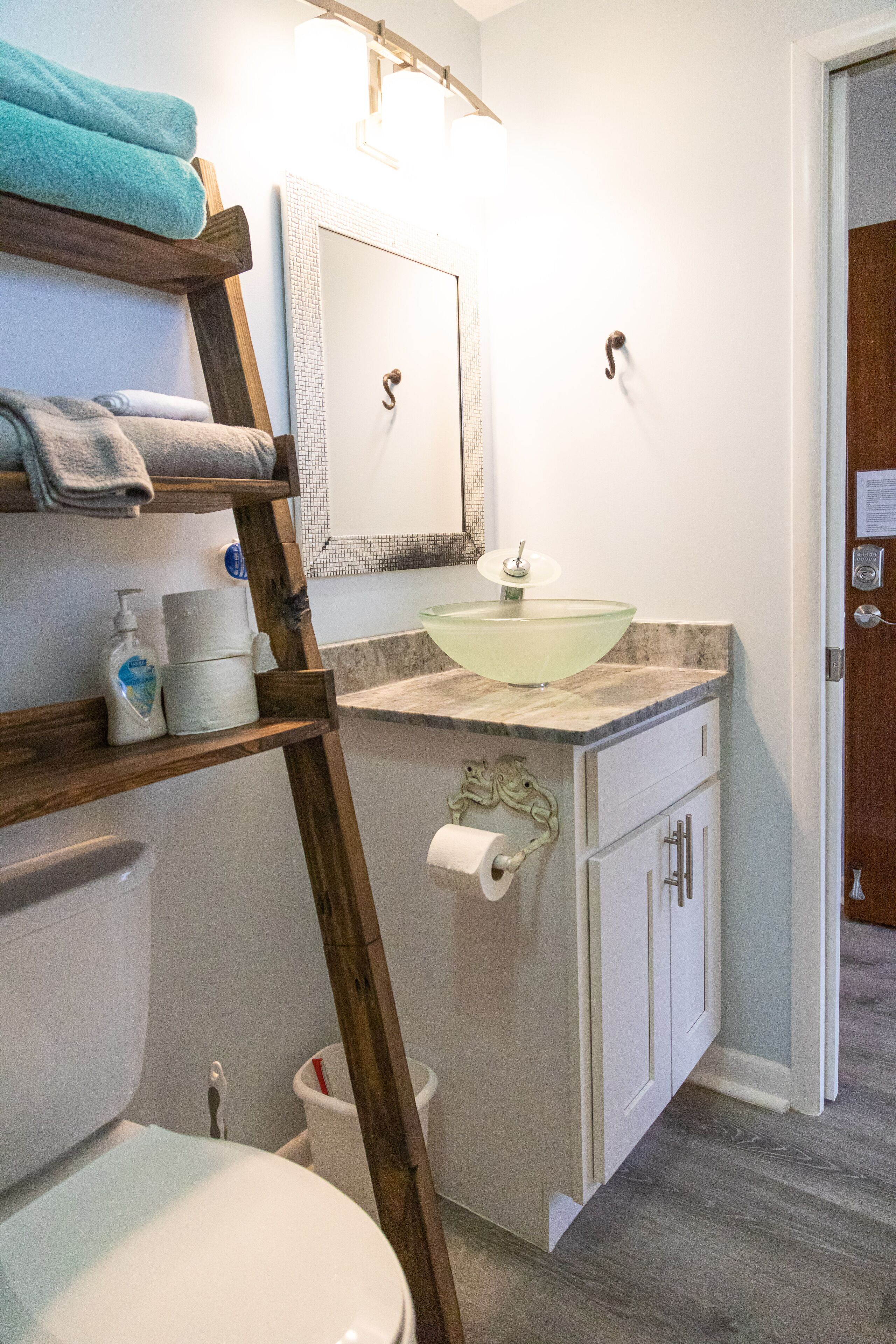 Une salle de bain comprenant des toilettes, un lavabo avec un récipient en verre, une étagère en bois avec des serviettes et un miroir.