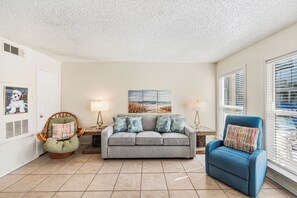 A living room with a grey sofa, a blue armchair, a wicker chair, two lamps, and a painting on the wall. - Poolside Escape | Private Patio & Steps to the Beach (Corpus Christi)