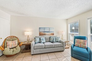 A living room with a grey sofa, a wicker chair, and a blue armchair. There are two lamps on side tables, a painting on the wall, and a door to the left. - Poolside Escape | Private Patio & Steps to the Beach (Corpus Christi)