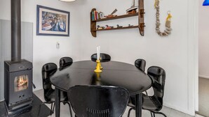 Un comedor con una mesa y sillas negras, un hogar a leña, una lámpara colgante y una estantería con libros y objetos decorativos.