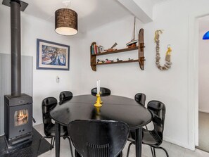 A dining area with a black table and chairs, a wood-burning stove, a hanging light fixture, and a bookshelf with books and decorative items.
