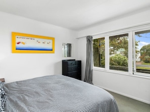 A bedroom with a bed, a dresser, a window with curtains, and a framed picture on the wall. - Wharewaka Bach - Lake Taupo Holiday Home (Wharewaka)