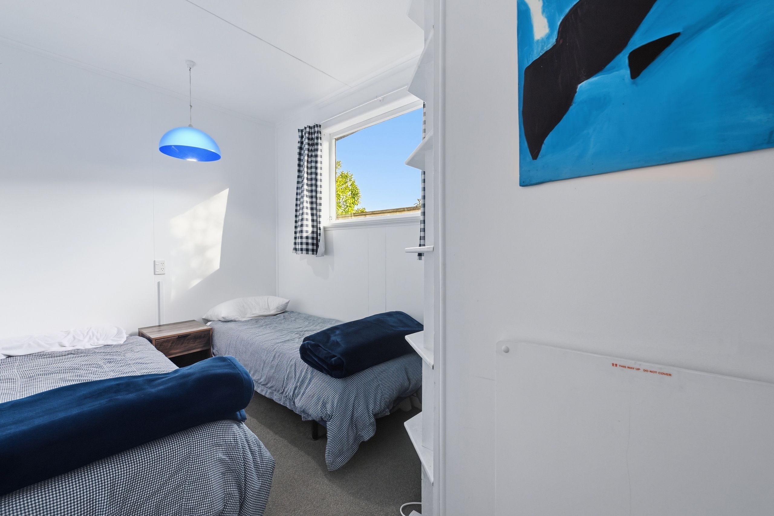 A small, well-lit room with two beds, a window with a view of greenery, and a blue artwork on the wall.