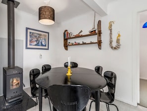 A dining area with a black table and chairs, a wood-burning stove, a hanging light fixture, and a bookshelf with books and decorative items. - Wharewaka Bach - Lake Taupo Holiday Home (Wharewaka)