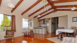 A spacious living area with a high ceiling, wooden beams, a dining table, and a kitchen in the background. - Views on Rimu - Whitianga Holiday Home (Whitianga)