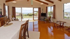 A spacious living room with a high ceiling, wooden beams, a dining area, and large windows offering a view of the outdoors. - Views on Rimu - Whitianga Holiday Home (Whitianga)