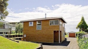 A small, single-story wooden house with a garage, surrounded by a grassy area and a paved driveway. - Views on Rimu - Whitianga Holiday Home (Whitianga)