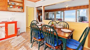 Un comedor con mesa y sillas de madera, una zona de cocina con un armario rojo, y vista al mar a través de amplios ventanales.