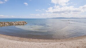 Una playa con agua cristalina, un rompeolas rocoso y montañas lejanas bajo un cielo azul.