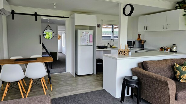 A modern kitchen with white cabinets, a central island, and a dining area with a wooden table and white chairs. - The reviews say it all! A blissful bach, & pet friendly! (Mangakino)