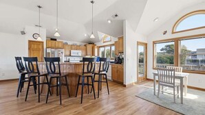 Een ruime keuken met houten kasten, een centraal eiland met stoelen, hanglampen en grote ramen met uitzicht op de omgeving.
