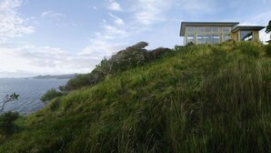 A modern building with large windows overlooking a grassy hillside and a body of water. - XSPOT Cliff 4WD holiday apartment for two - Spectacular 270° Hauraki Gulf views (Tryphena)