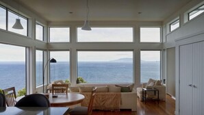 A spacious room with large windows offering a view of the sea. There is a dining table with chairs, a sofa, and a lamp. - XSPOT Cliff 4WD holiday apartment for two - Spectacular 270° Hauraki Gulf views (Tryphena)