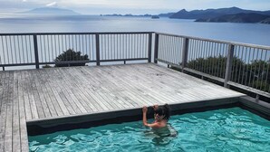A wooden deck with a swimming pool, a person swimming, and a view of the sea and mountains. - XSPOT Cliff 4WD holiday apartment for two - Spectacular 270° Hauraki Gulf views (Tryphena)