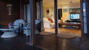 Une terrasse en bois avec des meubles en osier, un lit avec du linge de lit blanc et un téléviseur dans la pièce.