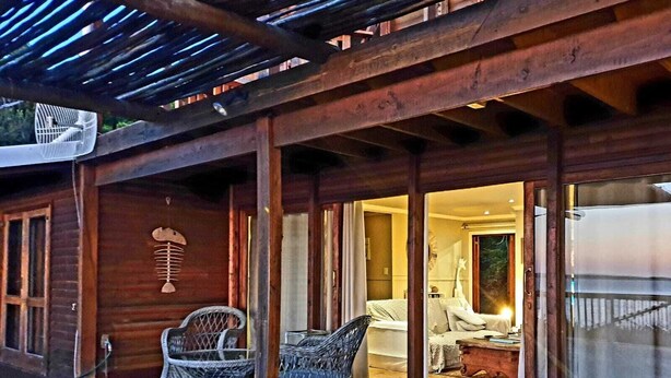 Une terrasse en bois, agrémentée de chaises et d’une table en osier, qui donne accès à une chambre avec un lit et une table de chevet.