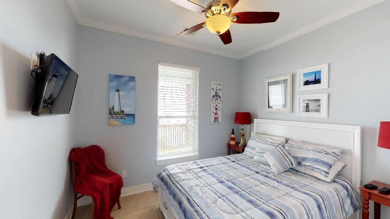 A bedroom with a bed, a ceiling fan, a TV mounted on the wall, a window with blinds, and a red chair.