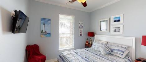 A bedroom with a bed, a ceiling fan, a TV mounted on the wall, a window with blinds, and a red chair.