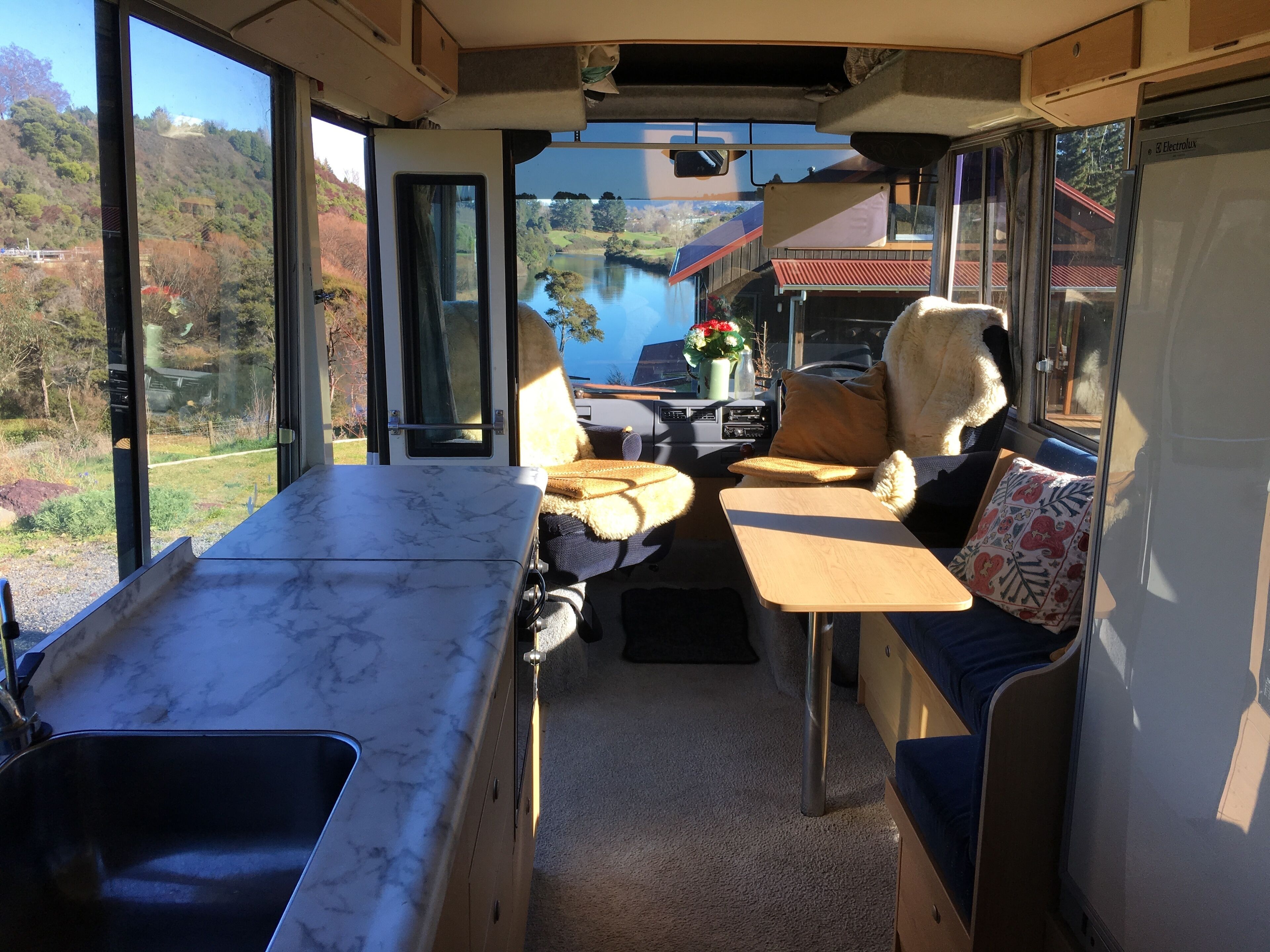 A camper van interior with a kitchen area, seating, and a view of a lake and hills.