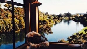 Een kamer met een groot raam dat uitkijkt op een rivier en groen. Er is een hondenbed met een pluizige hond, een klein tafeltje en een hangende lamp.