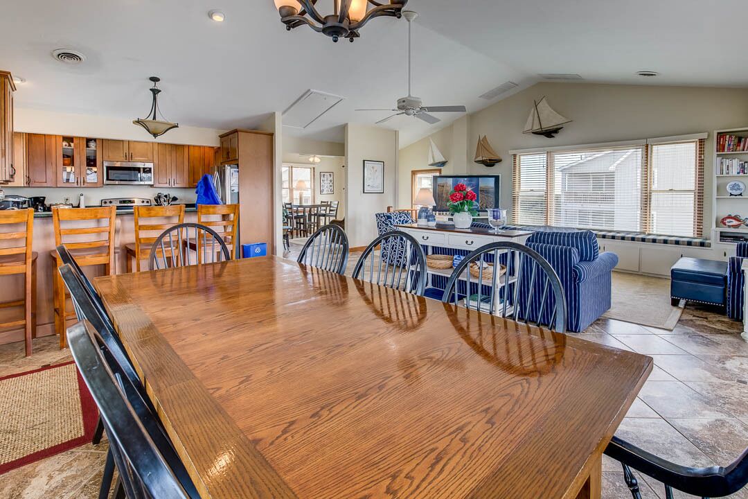 A spacious dining area with a wooden table and chairs, a kitchen with wooden cabinets, and a large window with blinds.