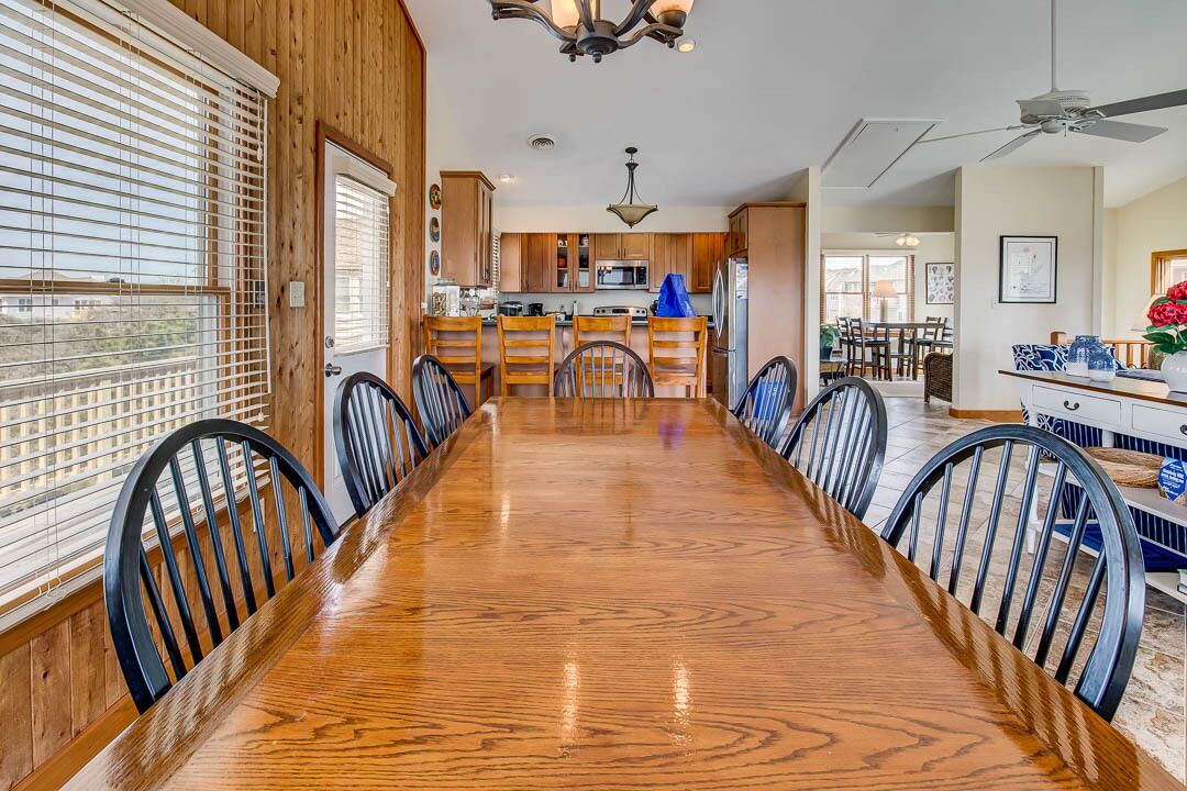 A dining area with a long wooden table and chairs, a kitchen in the background, and a ceiling fan.