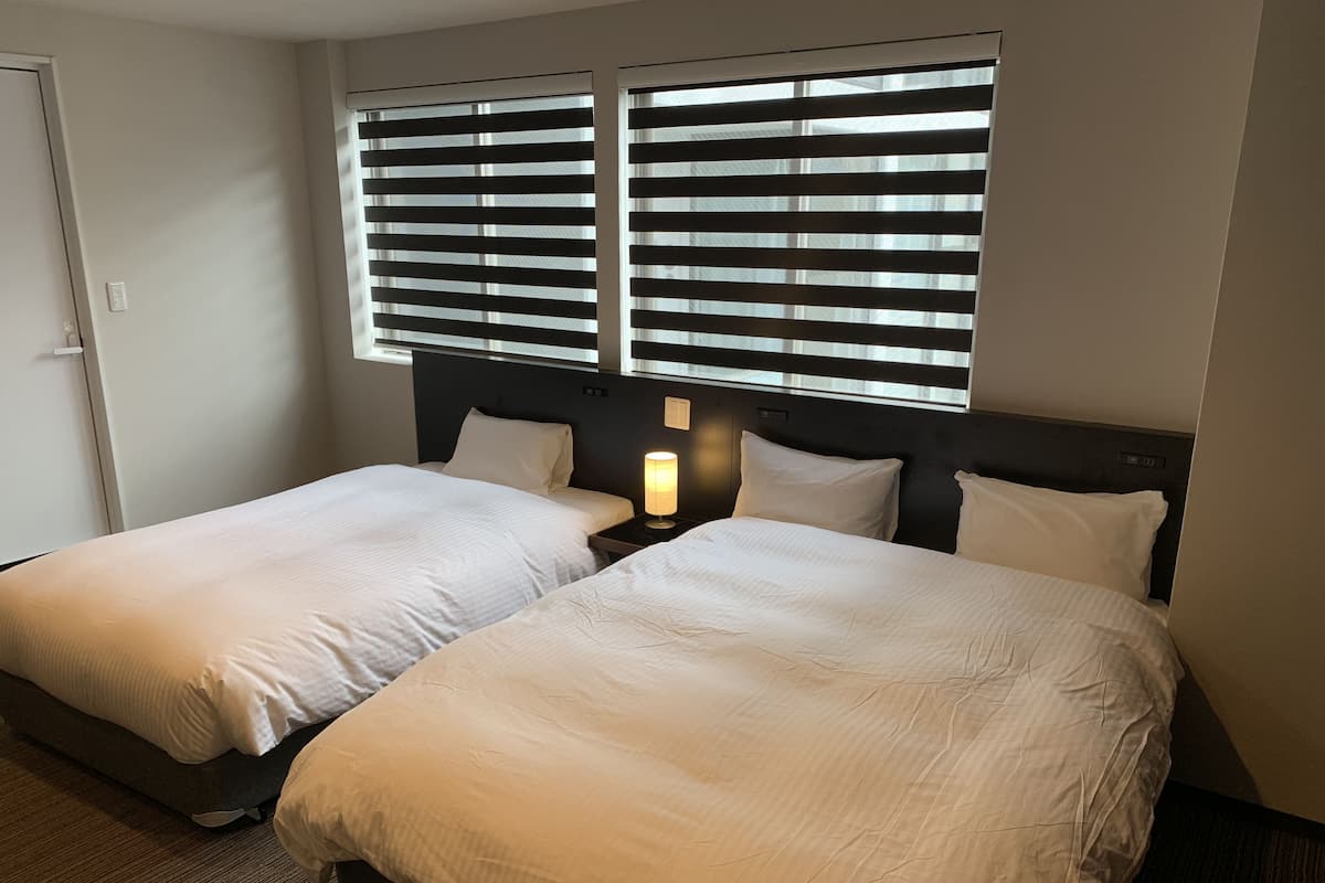 A hotel room with two beds, each with white bedding and a headboard. There are two windows with blinds, and a nightstand with a lamp between the beds.