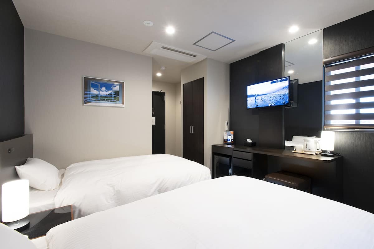 A hotel room with two beds, a TV mounted on the wall, a desk with a coffee maker, and a window with blinds.