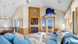 A cozy living room with a fireplace, blue sofas, and a large window overlooking a balcony. - Spur of the Moment | 293 ft from the beach | Private Pool, Hot Tub | Kill Devil Hills (Kill Devil Hills)