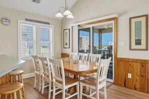Un comedor con una mesa de madera y sillas blancas, un reloj en la pared y vistas a través de un ventanal.