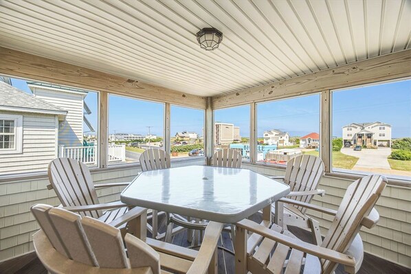 A covered outdoor dining area with a round table and six chairs, large windows offering a view of the surrounding neighborhood.