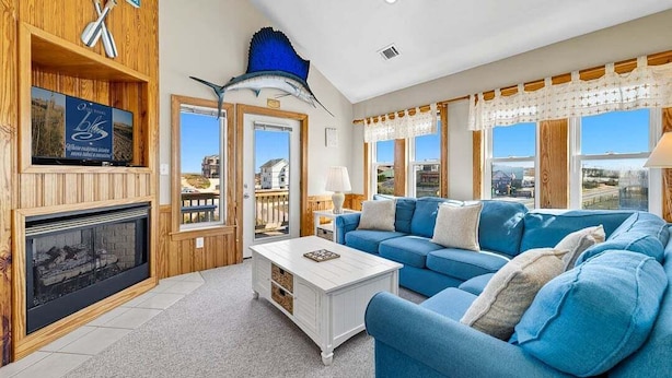 A spacious living room with a fireplace, a blue sofa, and a large window offering a view of the outside. - Spur of the Moment | 293 ft from the beach | Private Pool, Hot Tub | Kill Devil Hills (Kill Devil Hills)