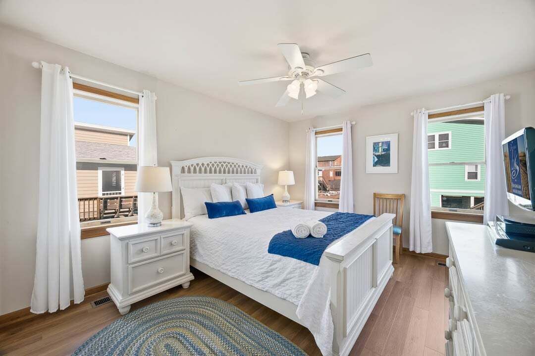 A bedroom with a white bed, blue pillows, a white nightstand, a ceiling fan, and a view of a building through the window.