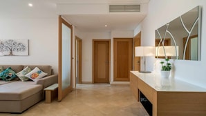 Una sala moderna con un sofá beige, una mesa de centro y una mesita auxiliar. Se ve una puerta de madera que da a otra habitación, un espejo en la pared y una planta en maceta sobre un aparador.