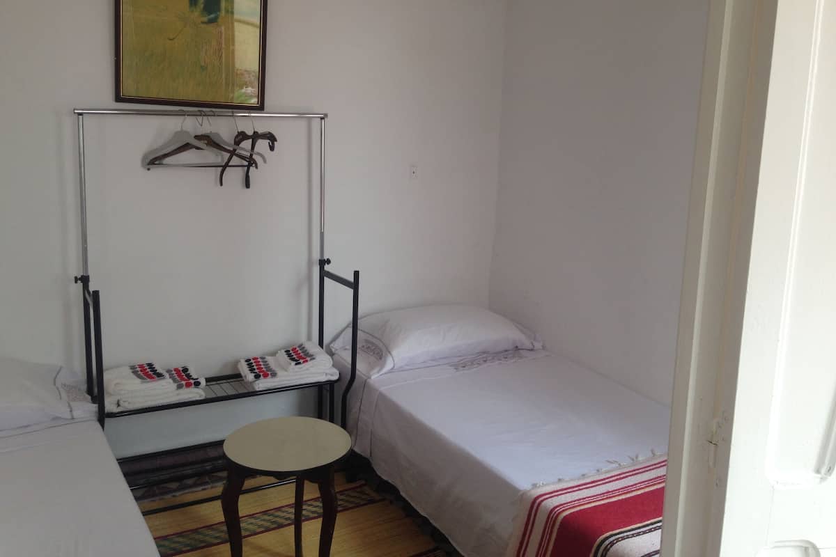 A small, simple room with a single bed, a small table, and a drying rack with towels.