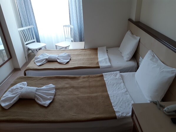 Quarto de hotel com duas camas, cada uma com um cobertor marrom e uma toalha branca amarrada em laço. Há uma pequena mesa e uma cadeira no canto, e uma janela com cortinas.