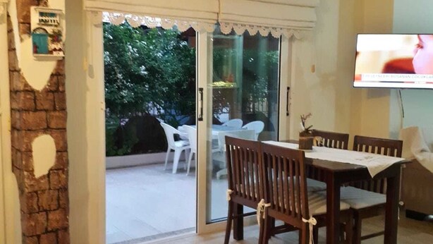 An interior with a dining area, a TV mounted on the wall, and a glass door leading to an outdoor space. - Ayhan Otel (Cesme)