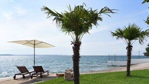 Escena costera con dos sillas de playa y una sombrilla grande, palmeras y vista al mar.