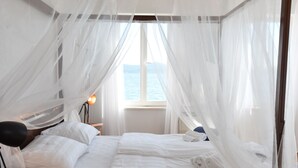 Un dormitorio con cama, cortinas blancas, una lámpara de noche y vista al mar.