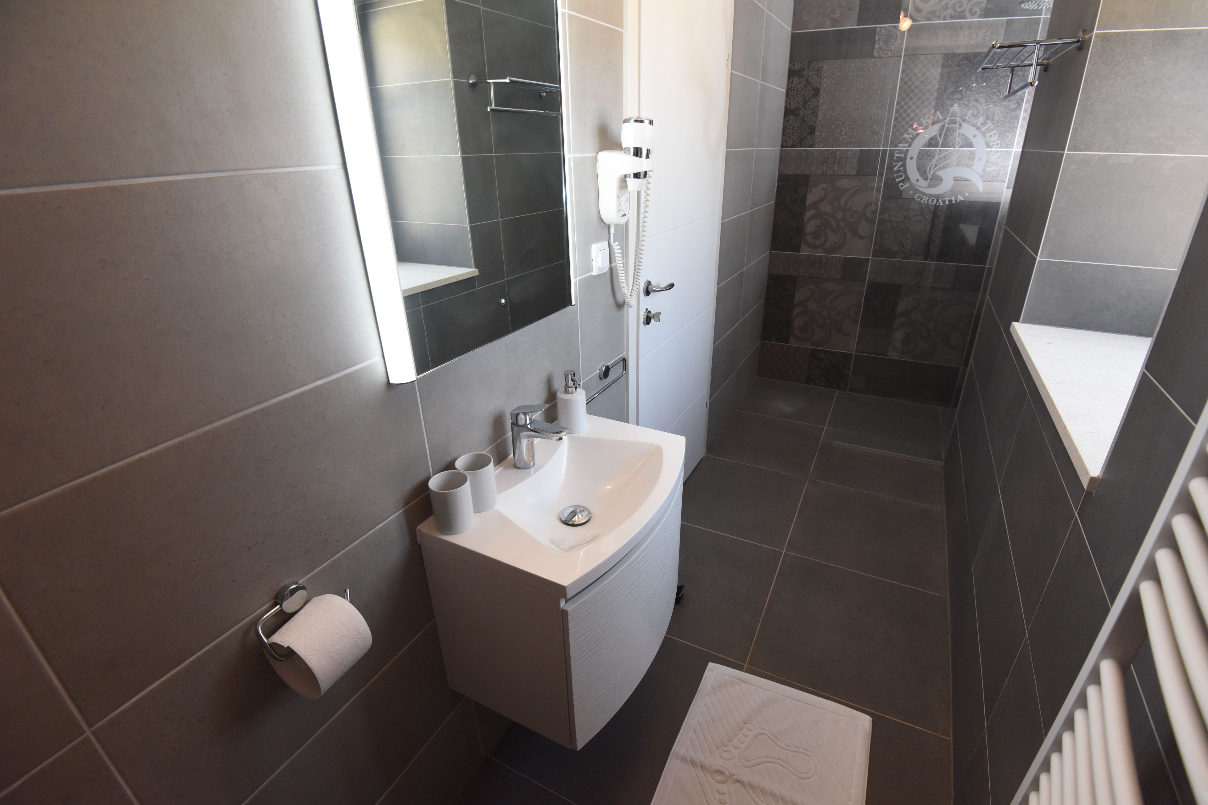 Un baño moderno con un lavamanos blanco, un espejo, una toalla y una zona de ducha.
