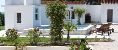 A white building with a swimming pool, a patio with lounge chairs, and a garden with various plants.