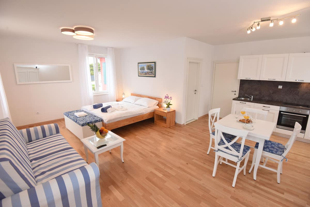 A modern, well-lit living room with a sofa, a dining table with chairs, a kitchen area, and a bed with a nightstand.