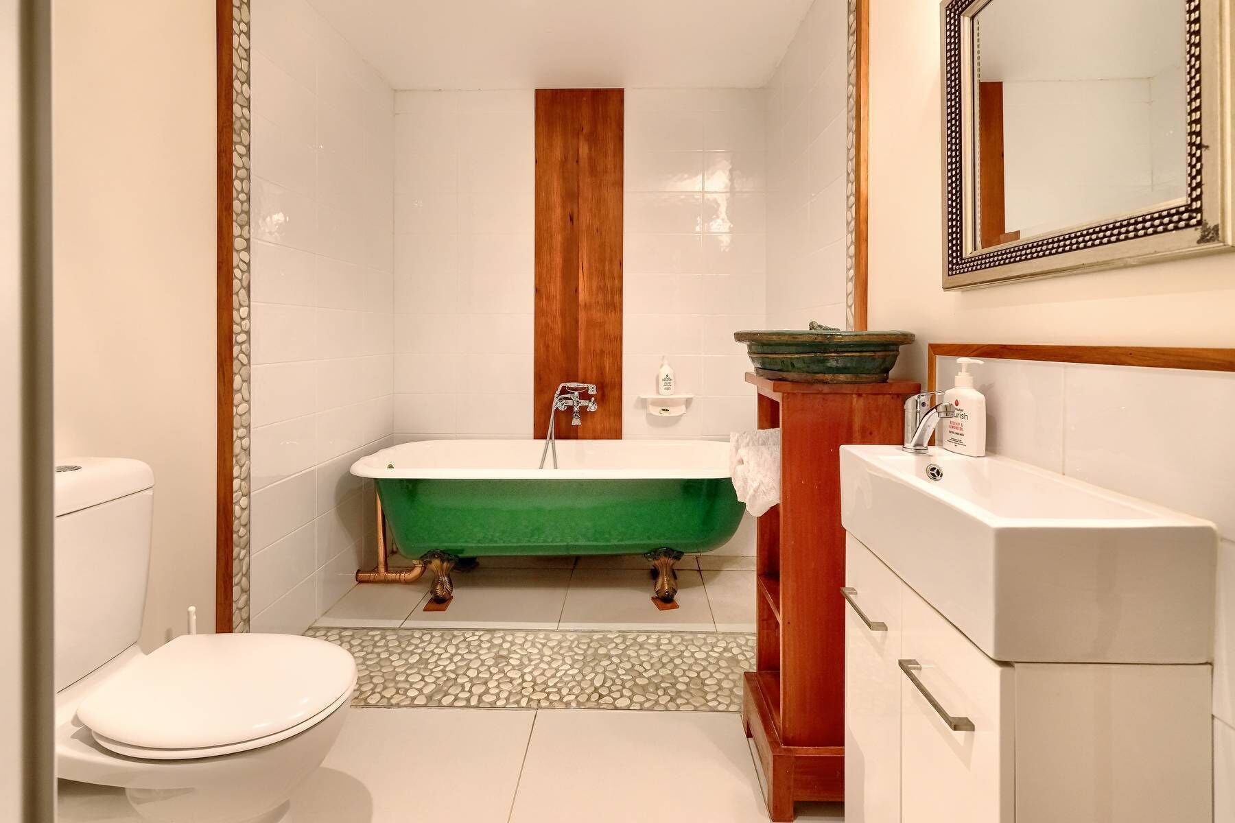 Casa de banho com uma banheira de patas de leão verde, sanita branca e toucador de madeira com espelho.