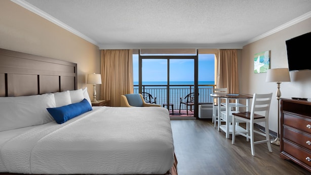Ein Hotelzimmer mit einem großen Bett, einem Esstisch, Stühlen, einem Fernseher und einem Balkon mit Meerblick.