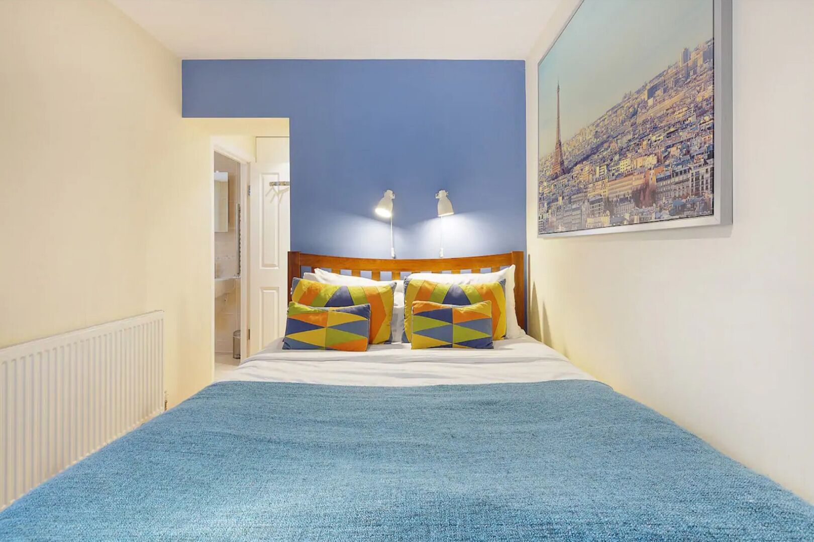 A bedroom with a bed, colorful pillows, a blue blanket, a headboard, and a cityscape painting on the wall.